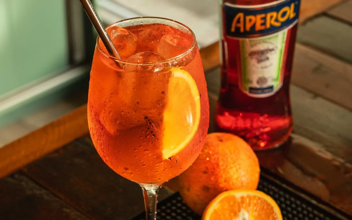 Copo com bebida alaranjada, gelo e fatia de laranja, acompanhado de garrafa de Aperol e laranjas frescas em bandeja preta