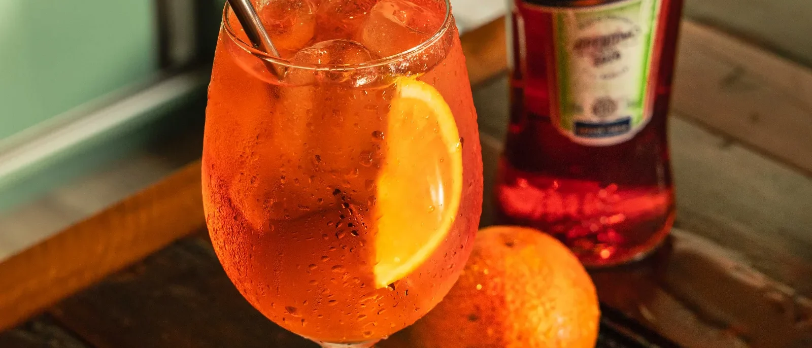 Copo com bebida alaranjada, gelo e fatia de laranja, acompanhado de garrafa de Aperol e laranjas frescas em bandeja preta