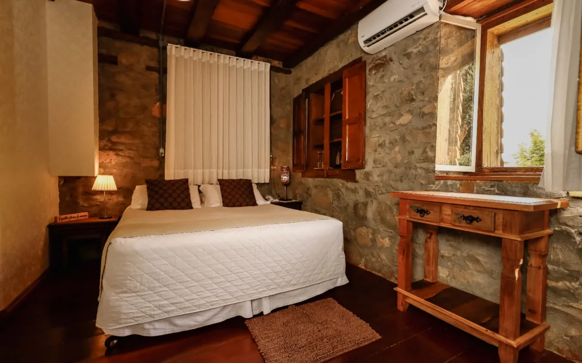 Quarto com cama de casal, paredes de pedra, móveis de madeira, janela com cortina branca, ar-condicionado e iluminação aconchegante