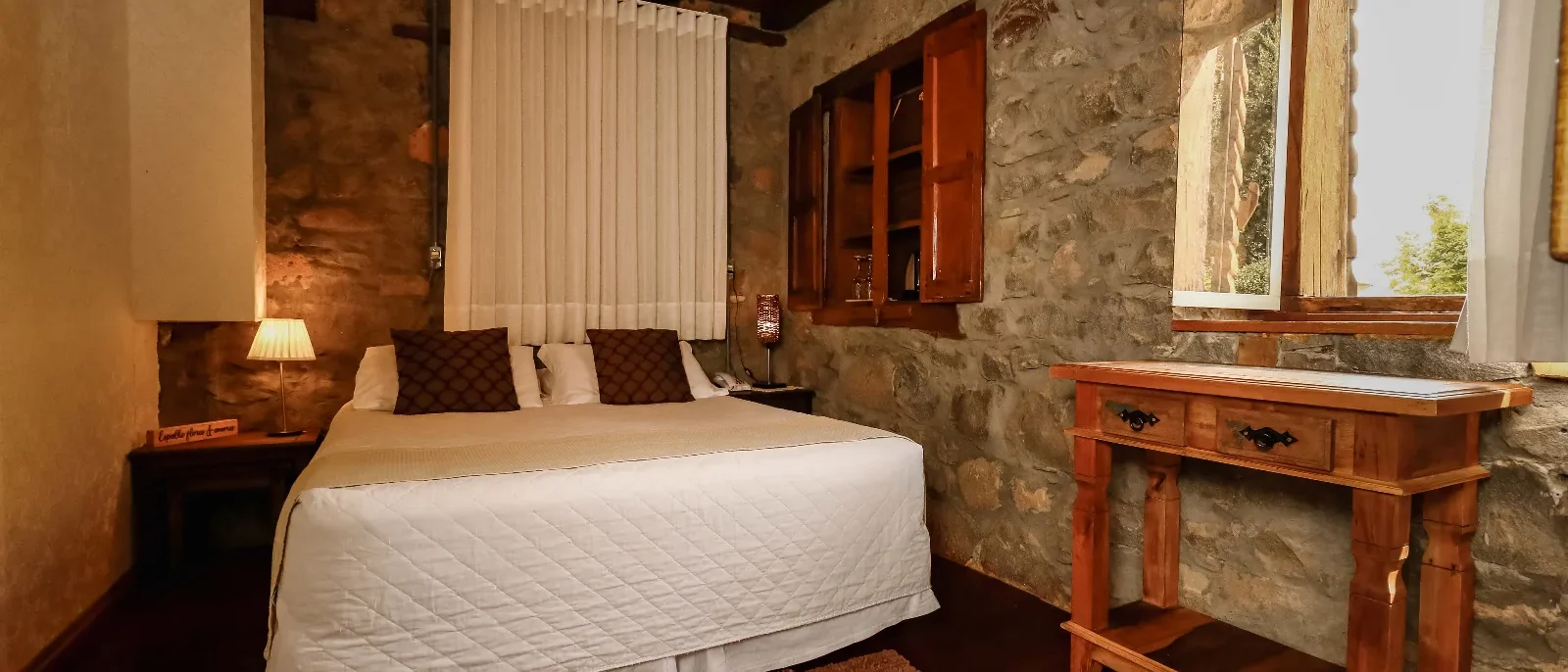 Quarto com cama de casal, paredes de pedra, móveis de madeira, janela com cortina branca, ar-condicionado e iluminação aconchegante