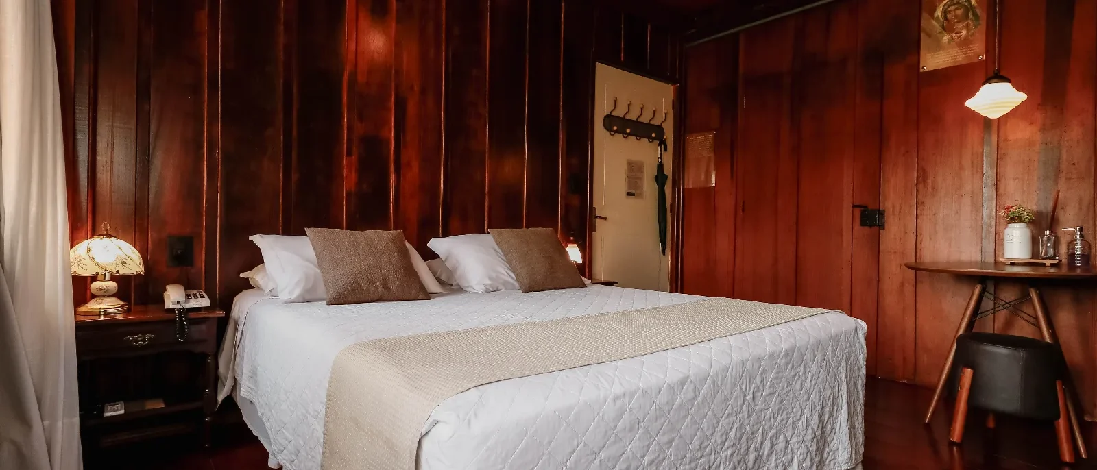 Quarto elegante com paredes de madeira avermelhada, cama de casal com roupa de cama branca e almofadas bege, criado com luminária, porta e decorações