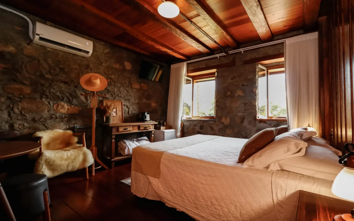 Quarto rústico com paredes de pedra, teto de madeira, cama com roupa de cama bege, janelas amplas e móveis de madeira clara
