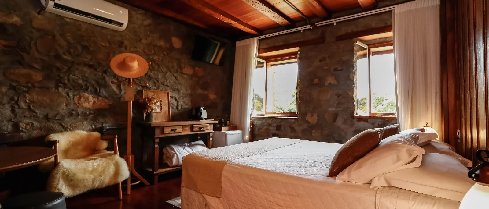 Quarto rústico com paredes de pedra, teto de madeira, cama com roupa de cama bege, janelas amplas e móveis de madeira clara