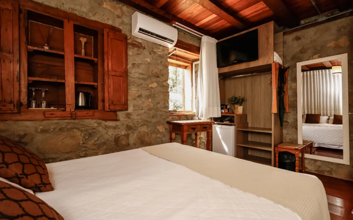 Quarto com cama de casal, paredes de concreto aparente, móveis de madeira, janela com cortina branca, ar-condicionado e TV de parede