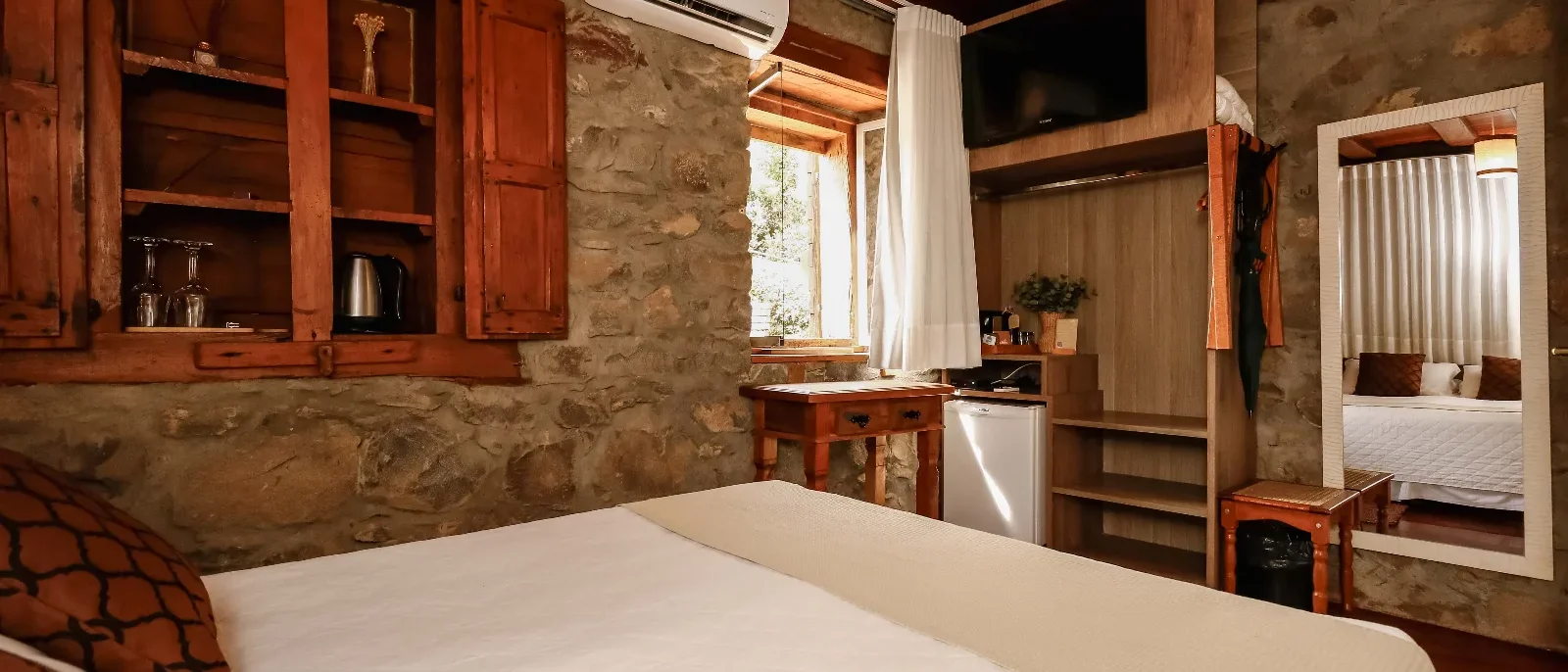 Quarto com cama de casal, paredes de concreto aparente, móveis de madeira, janela com cortina branca, ar-condicionado e TV de parede