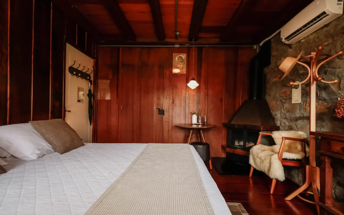 Quarto com paredes de madeira avermelhada, cama de casal com roupa de cama branca, lareira, cadeiras de madeira com pele de cordeiro e objetos decorativos