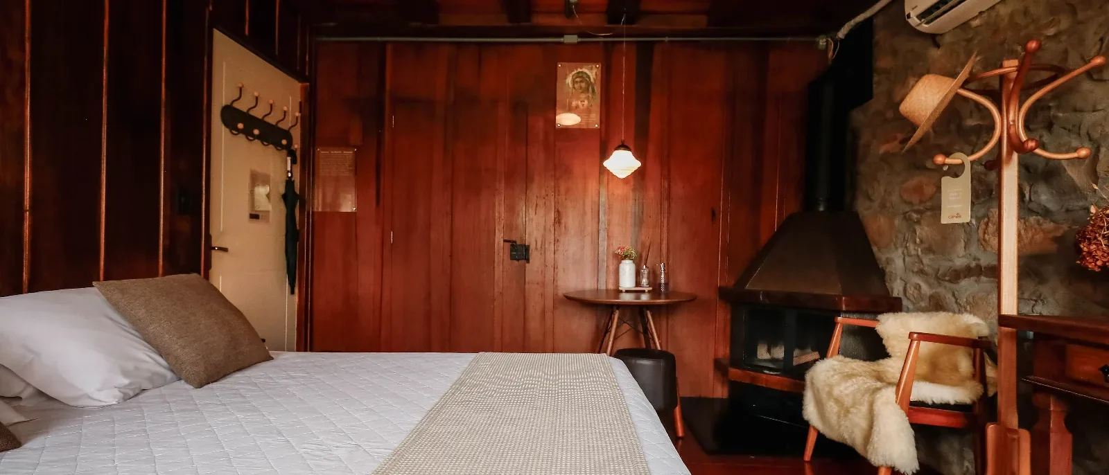 Quarto com paredes de madeira avermelhada, cama de casal com roupa de cama branca, lareira, cadeiras de madeira com pele de cordeiro e objetos decorativos