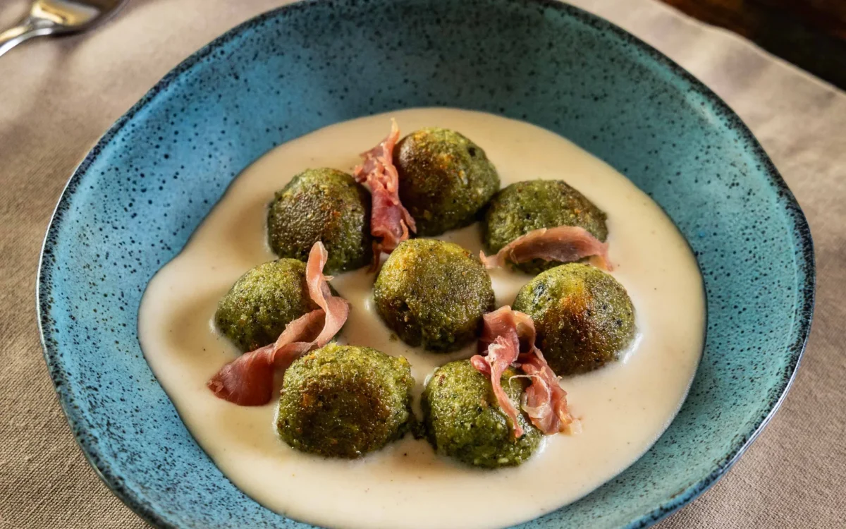 Prato azul com bolinhas verdes cobertas de tempero, creme bege, fatias de presunto rosa, dois garfos prateados e guardanapo bege sobre mesa de madeira