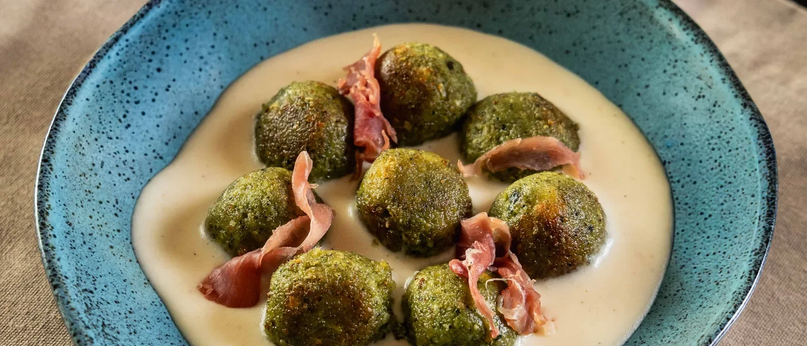 Prato azul com bolinhas verdes cobertas de tempero, creme bege, fatias de presunto rosa, dois garfos prateados e guardanapo bege sobre mesa de madeira