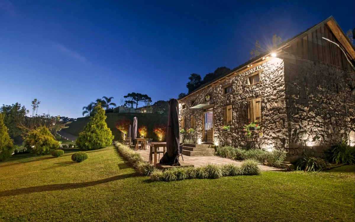 Casa de pedra iluminada ao entardecer com jardim bem cuidado, árvores e pátio com móveis, sob céu azul estrelado