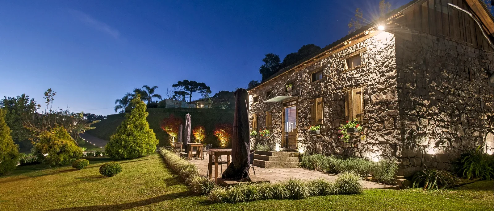 Casa de pedra iluminada ao entardecer com jardim bem cuidado, árvores e pátio com móveis, sob céu azul estrelado