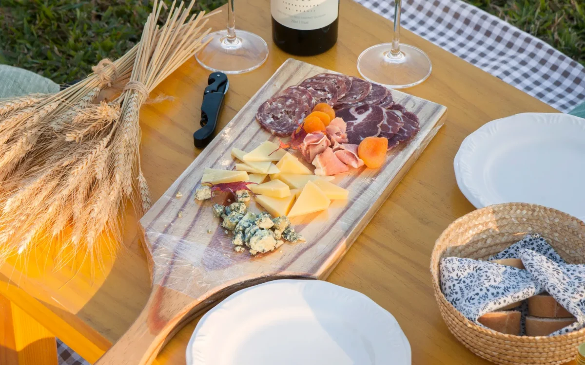 Mesa de piquenique com vinho, taças, charcutaria, queijos, pão, pratos brancos e decoração rústica sobre fundo de grama verde