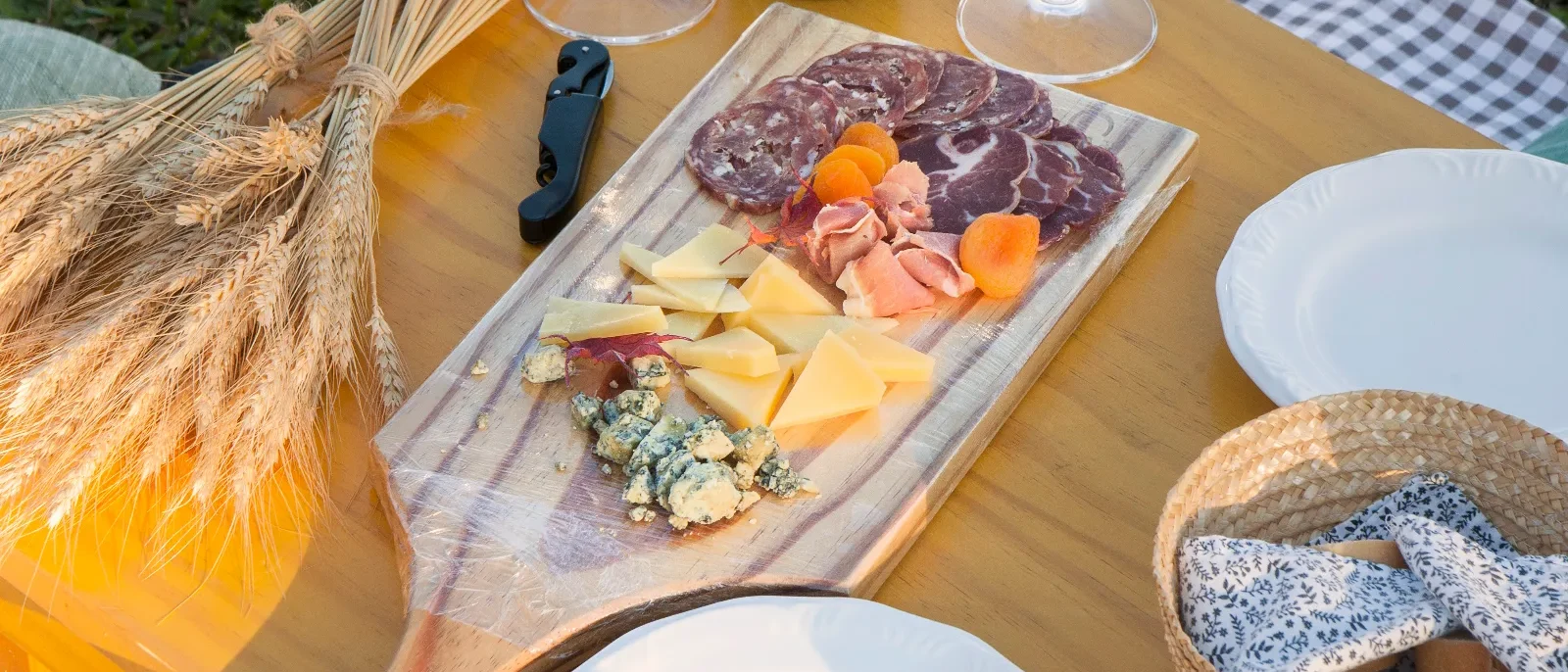 Mesa de piquenique com vinho, taças, charcutaria, queijos, pão, pratos brancos e decoração rústica sobre fundo de grama verde