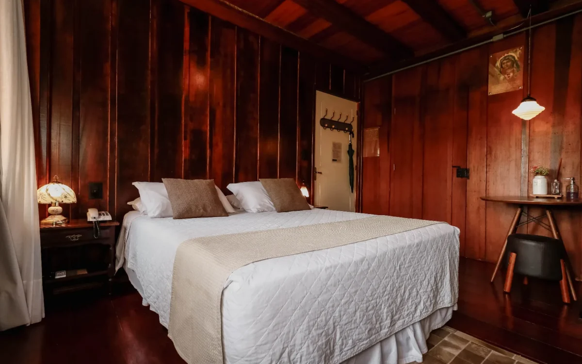 Quarto elegante com paredes de madeira avermelhada, cama de casal com roupa de cama branca e almofadas bege, criado com luminária, porta e decorações
