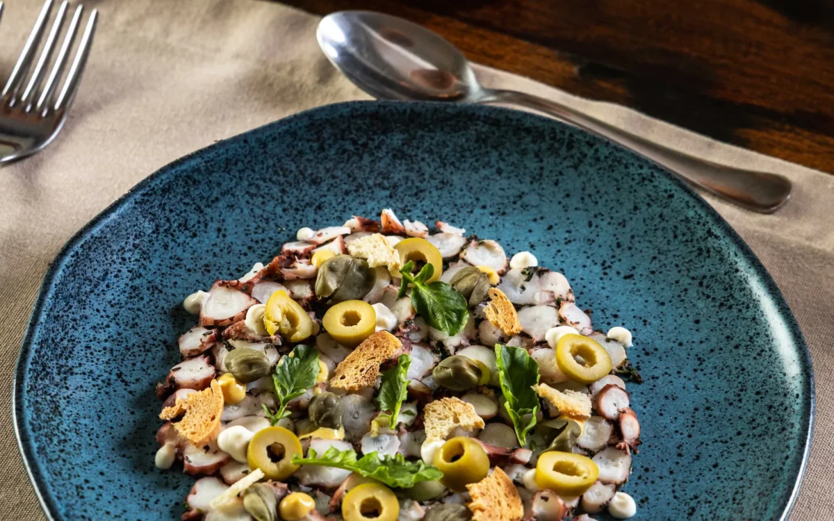 Prato azul com salada de frutos do mar, azeitonas verdes, camarões, camarão e gergelim, acompanhado de pão tostado e talheres numa mesa de madeira