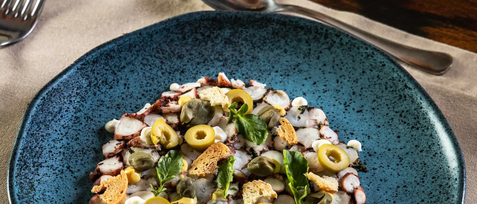 Prato azul com salada de frutos do mar, azeitonas verdes, camarões, camarão e gergelim, acompanhado de pão tostado e talheres numa mesa de madeira