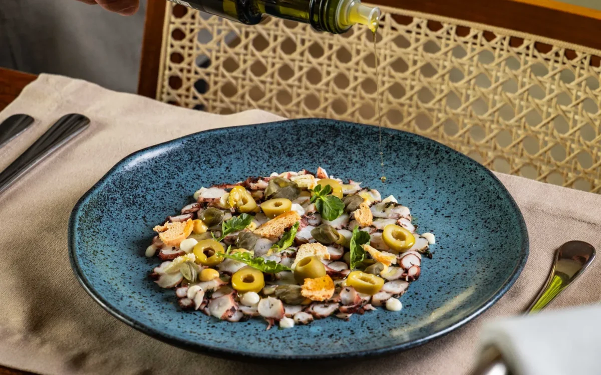 Mão de pessoa despejando azeite de oliva em prato azul com salada de frutos do mar, azeitonas e ervas frescas em mesa de madeira