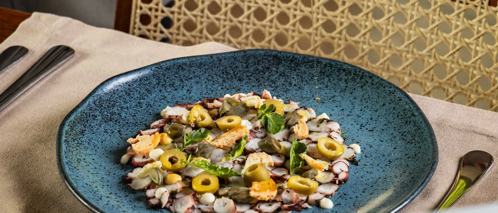 Mão de pessoa despejando azeite de oliva em prato azul com salada de frutos do mar, azeitonas e ervas frescas em mesa de madeira